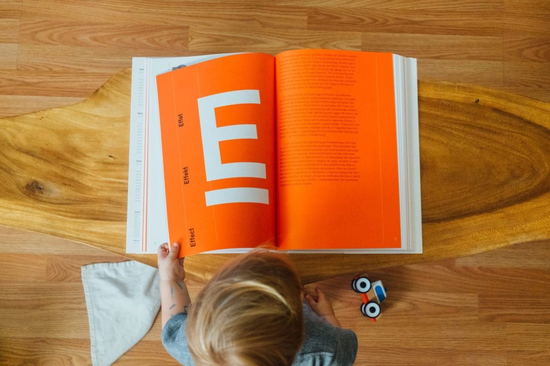 Child sitting on hardwood floor reading book with orange ‘E’ on left page from above.