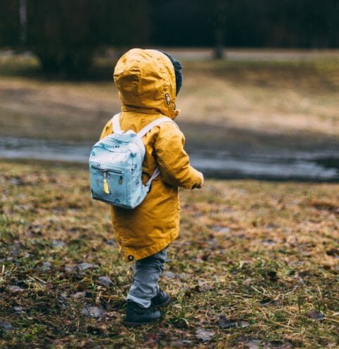 Child in yellow raincoat with backpack walking through grass field.