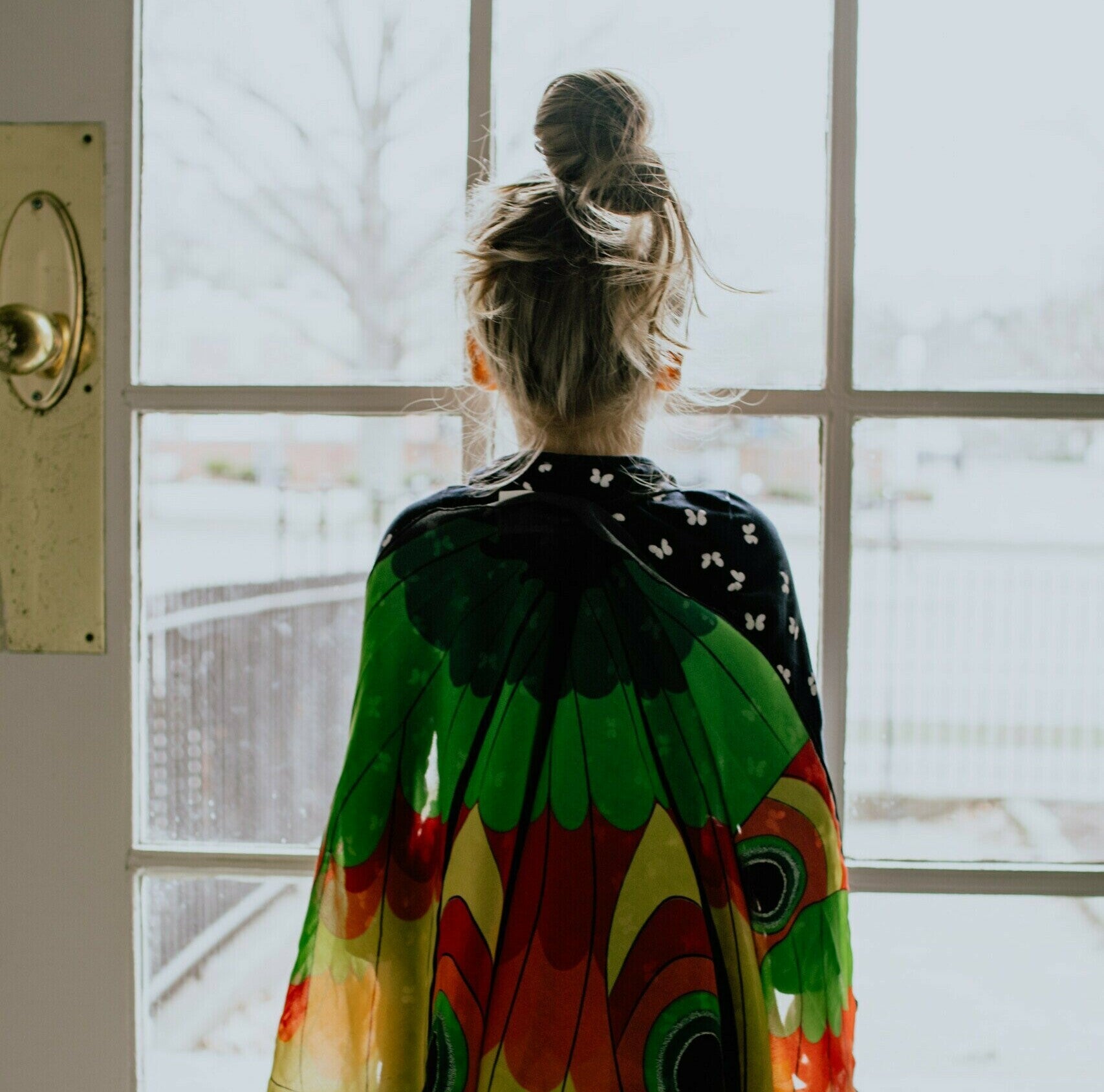 Young girl in multi-colored cape looking out a door.